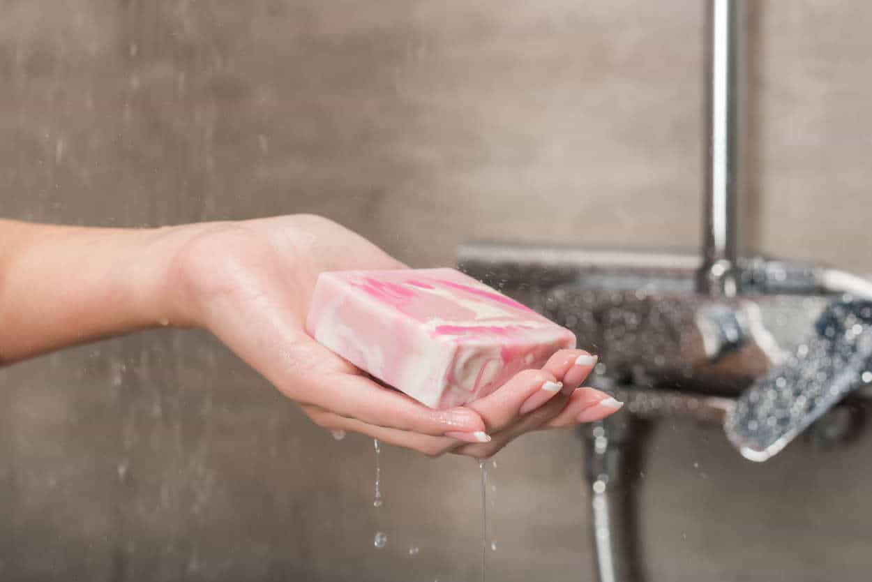 Close up image of a woman holding a bar of pink soap in her shower. The shower has no white streaks from hard water build up due to the installation of a water softener in Chandler Arizona