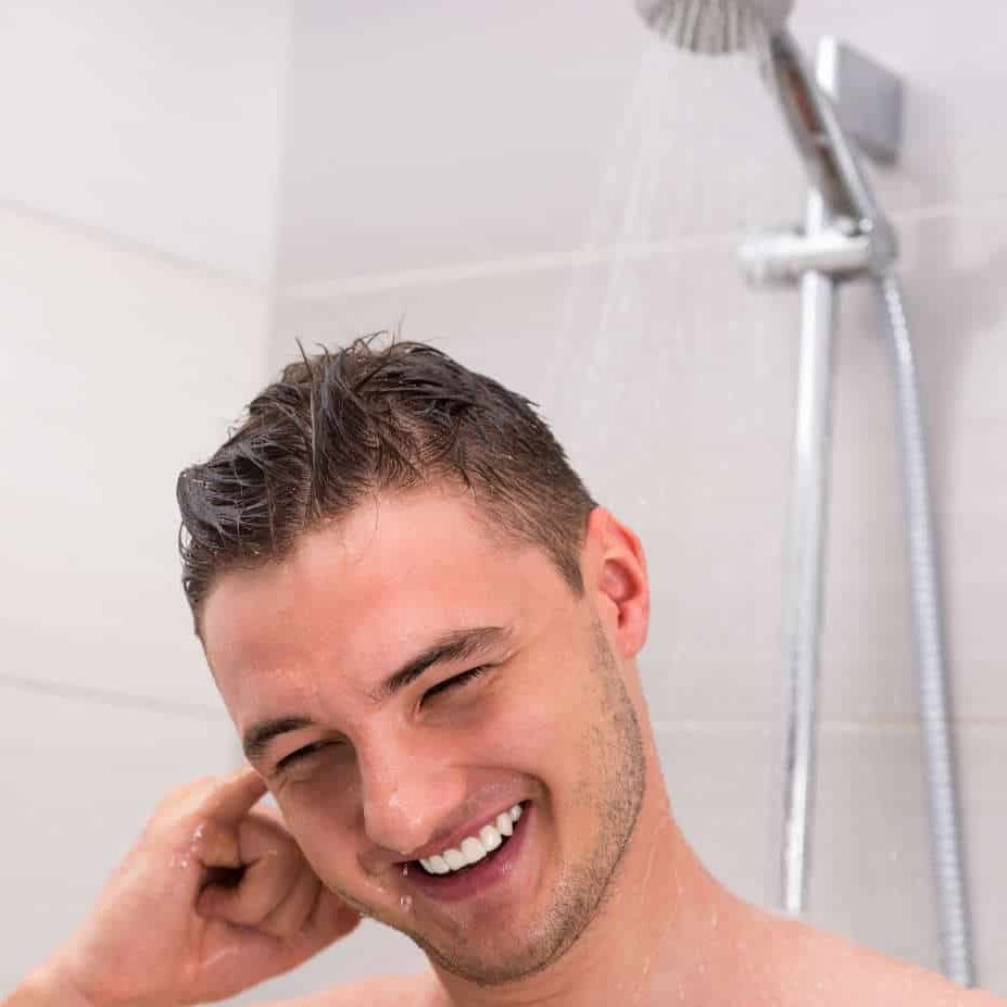 Young man cleaning his ear while taking a shower and standing under flowing water in the modern tiled bathroom
